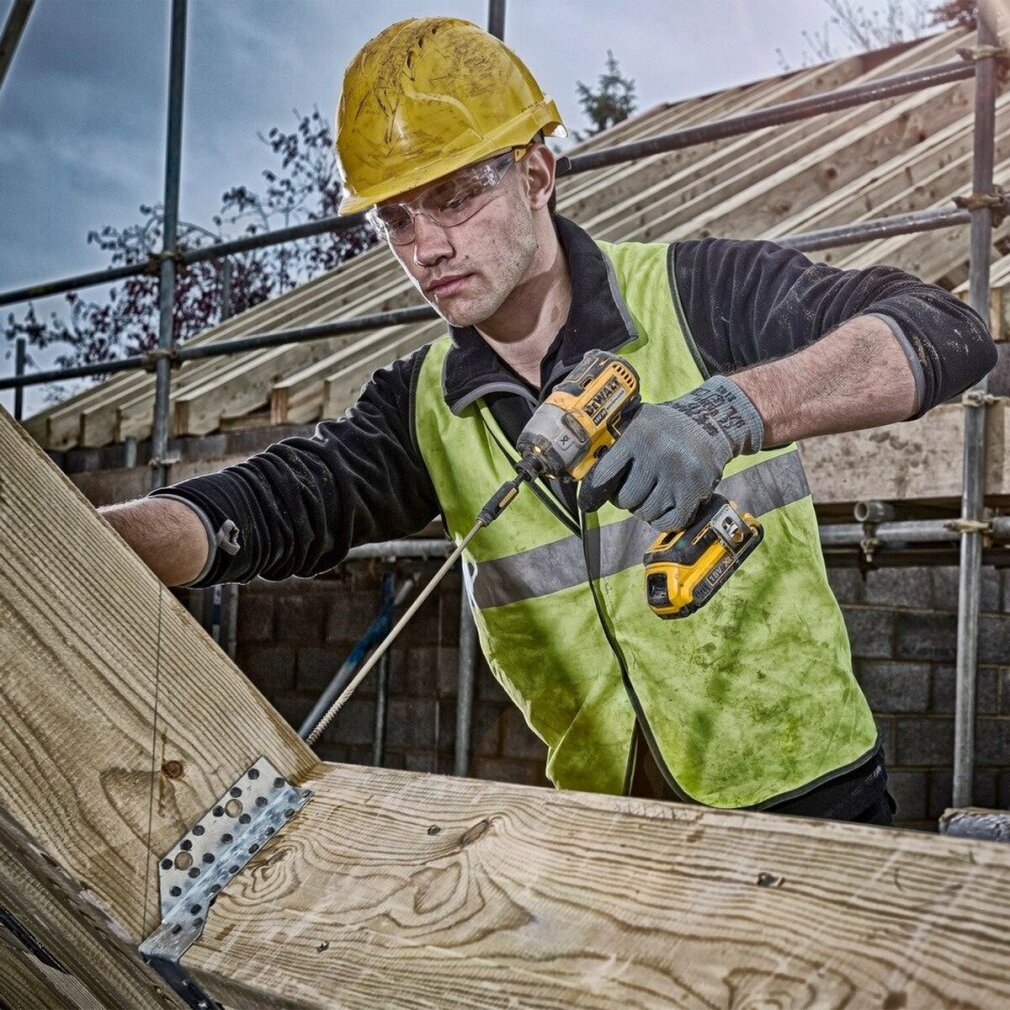 Robot budowlany w sprzęcie ochronnym używa wiertarki do drewna. Ma żółty kask, okulary i zieloną kamizelkę.