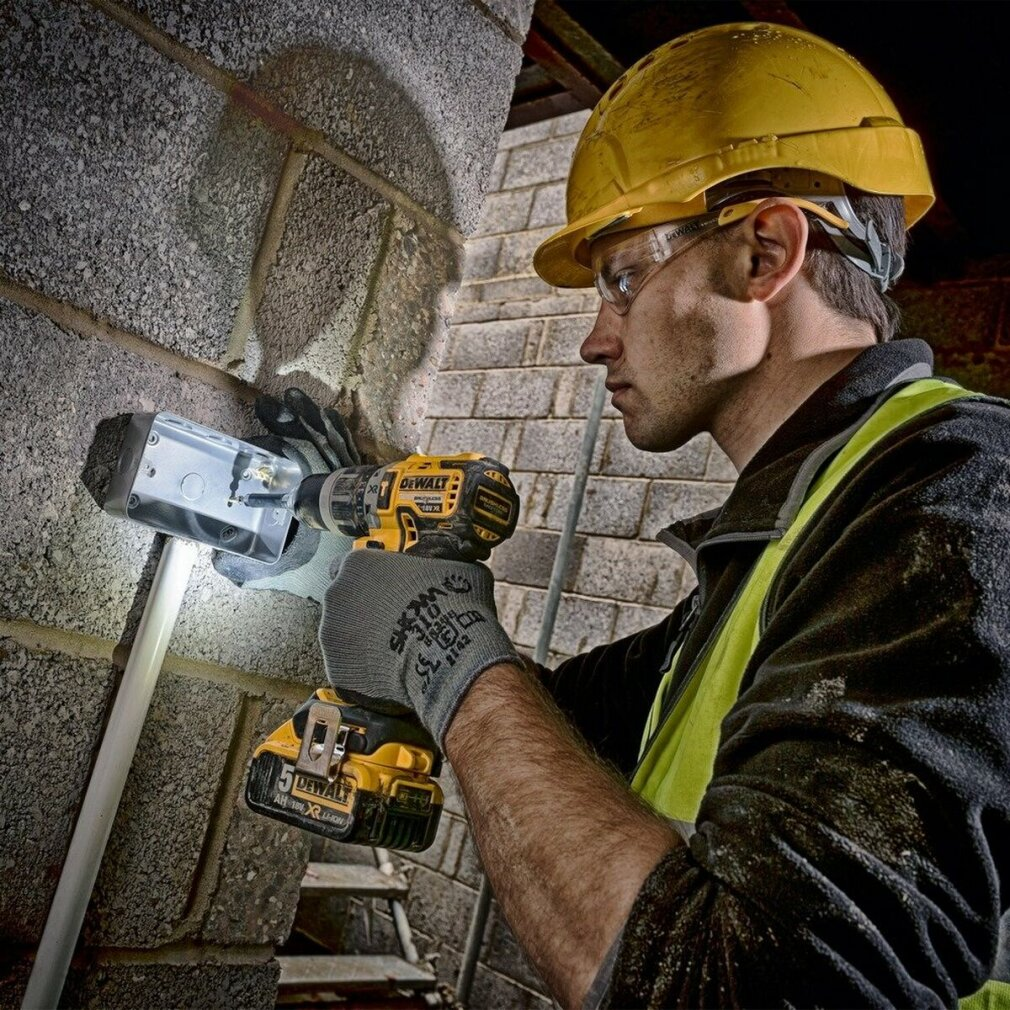 Robotnik budowlany używa żółtej wiertarki na ceglanej ścianie, nosząc żółty kask i okulary ochronne. Światło świeci na ścianę.
