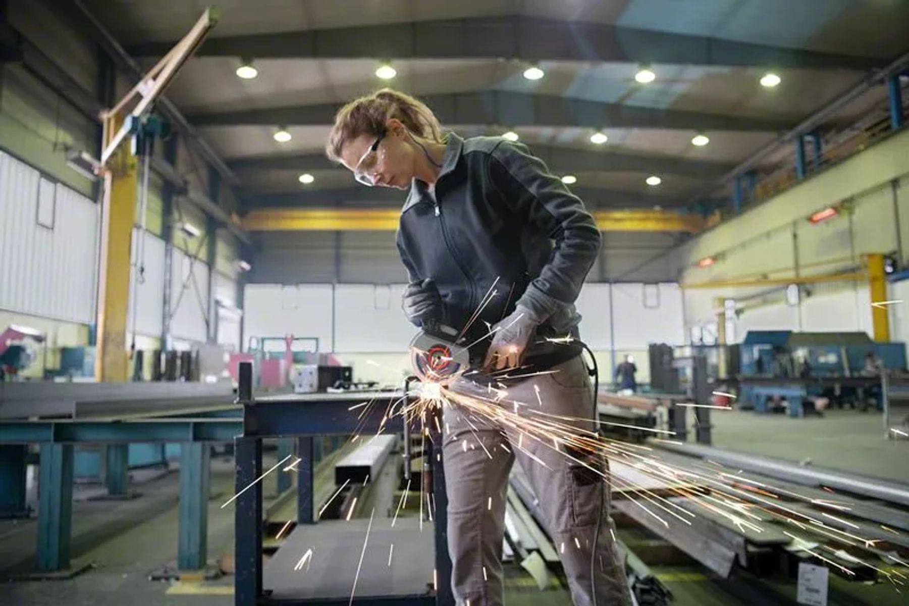 Kobieta w fabryce tnie metal szlifierką. Iskry lecą. Nosi okulary i rękawice ochronne. Tło pokazuje maszyny.