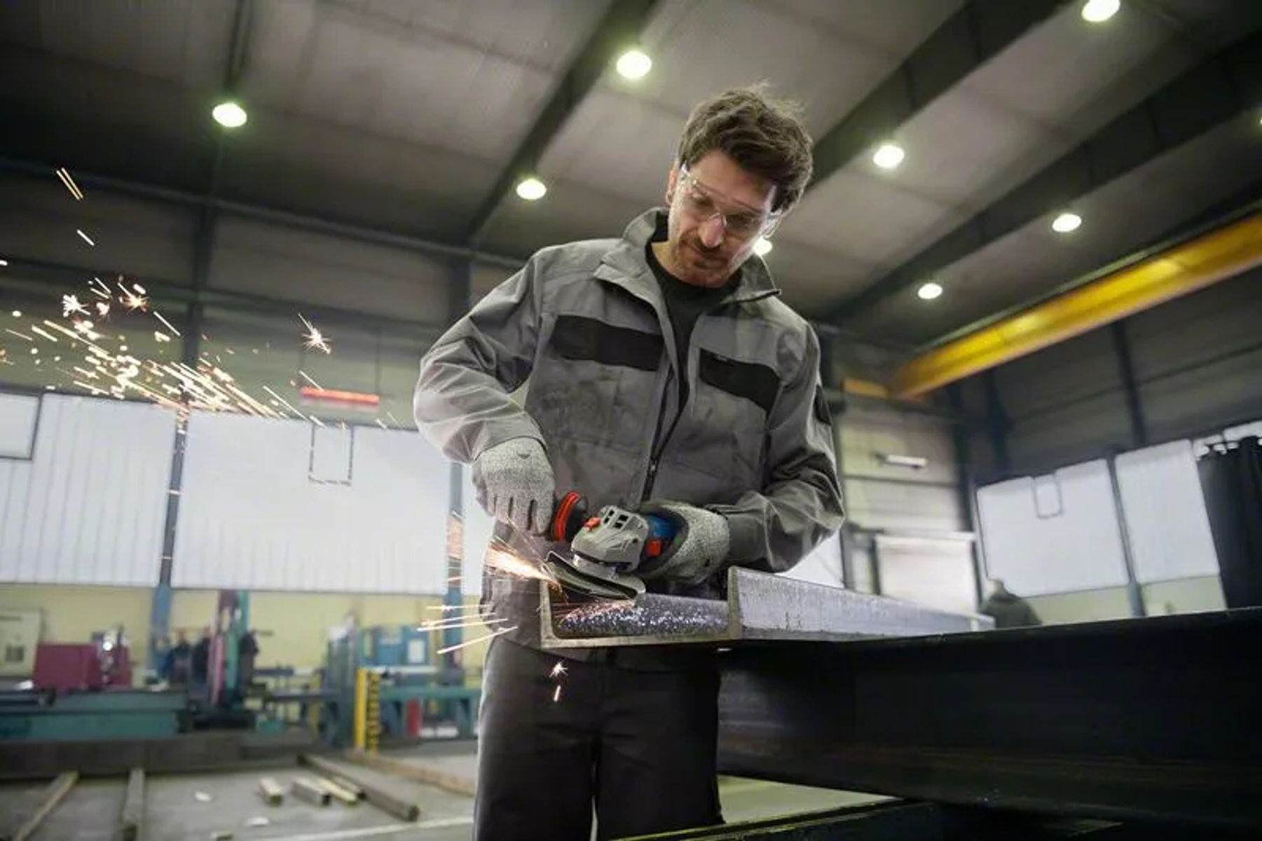 Mężczyzna w okularach ochronnych szlifuje metal, lecą iskry. Ma na sobie szary kombinezon roboczy i rękawice. Scena to warsztat.