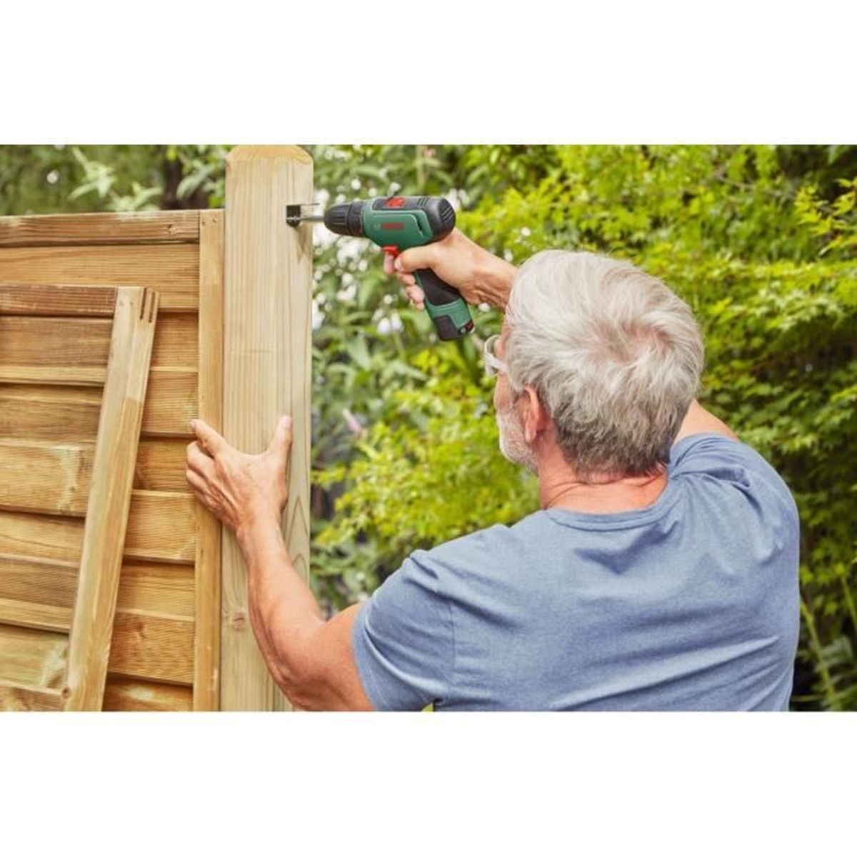 Mężczyzna używa wiertarki do montażu drewnianego płotu. Ma na sobie niebieską koszulę i pracuje na zewnątrz. Tło jest zielone.
