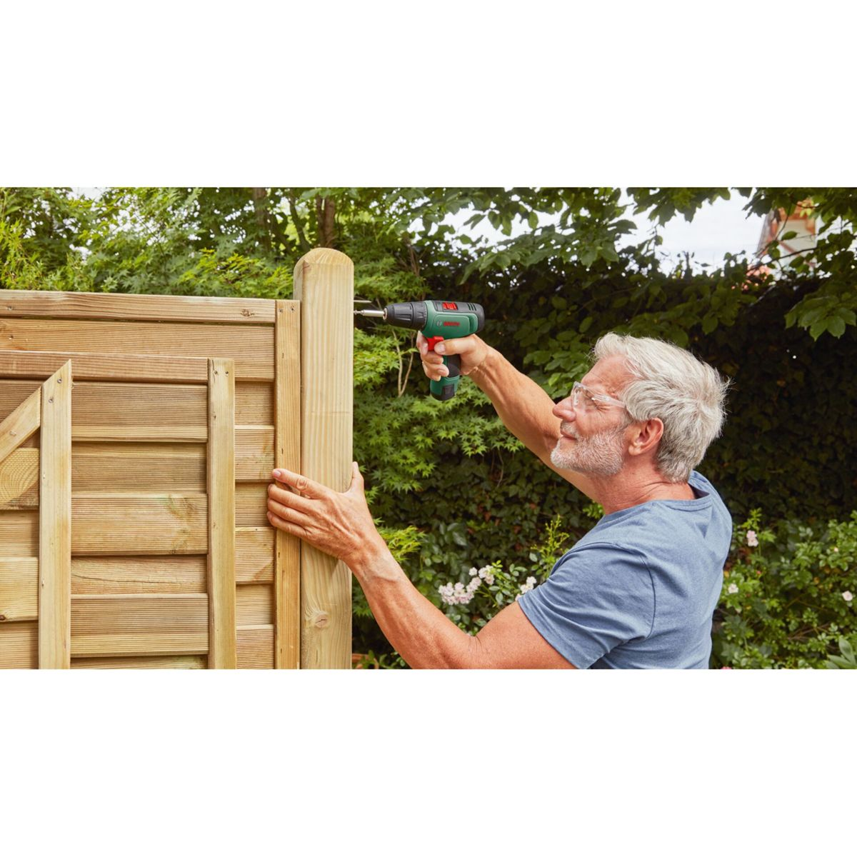 Mężczyzna wkręca śrubę w drewniany słupek ogrodzenia wiertarką. Ma okulary i niebieską koszulę.