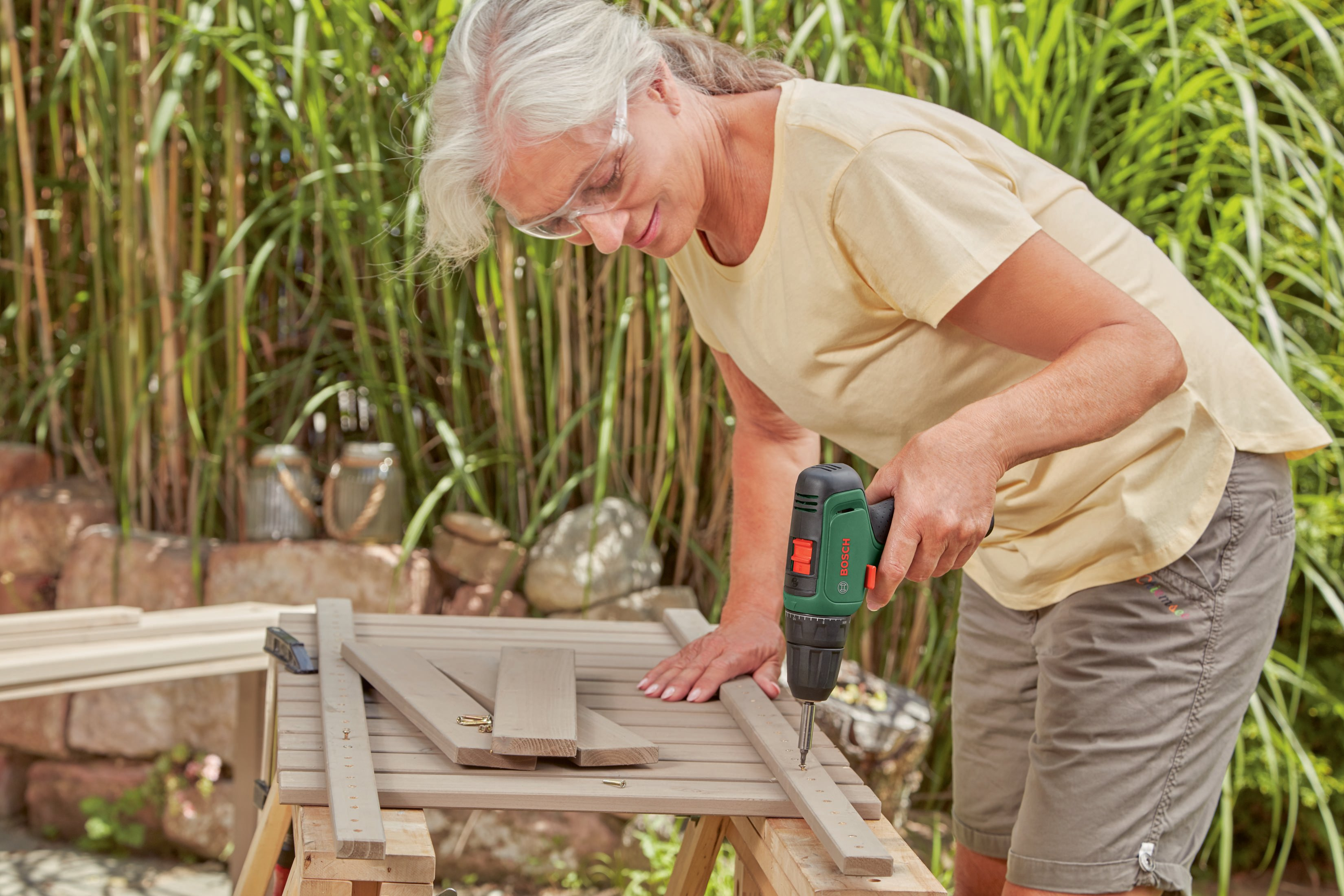 Kobieta wierci drewnianą deskę wiertarką elektryczną. Nosi okulary ochronne i żółtą koszulę, pracuje na zewnątrz.