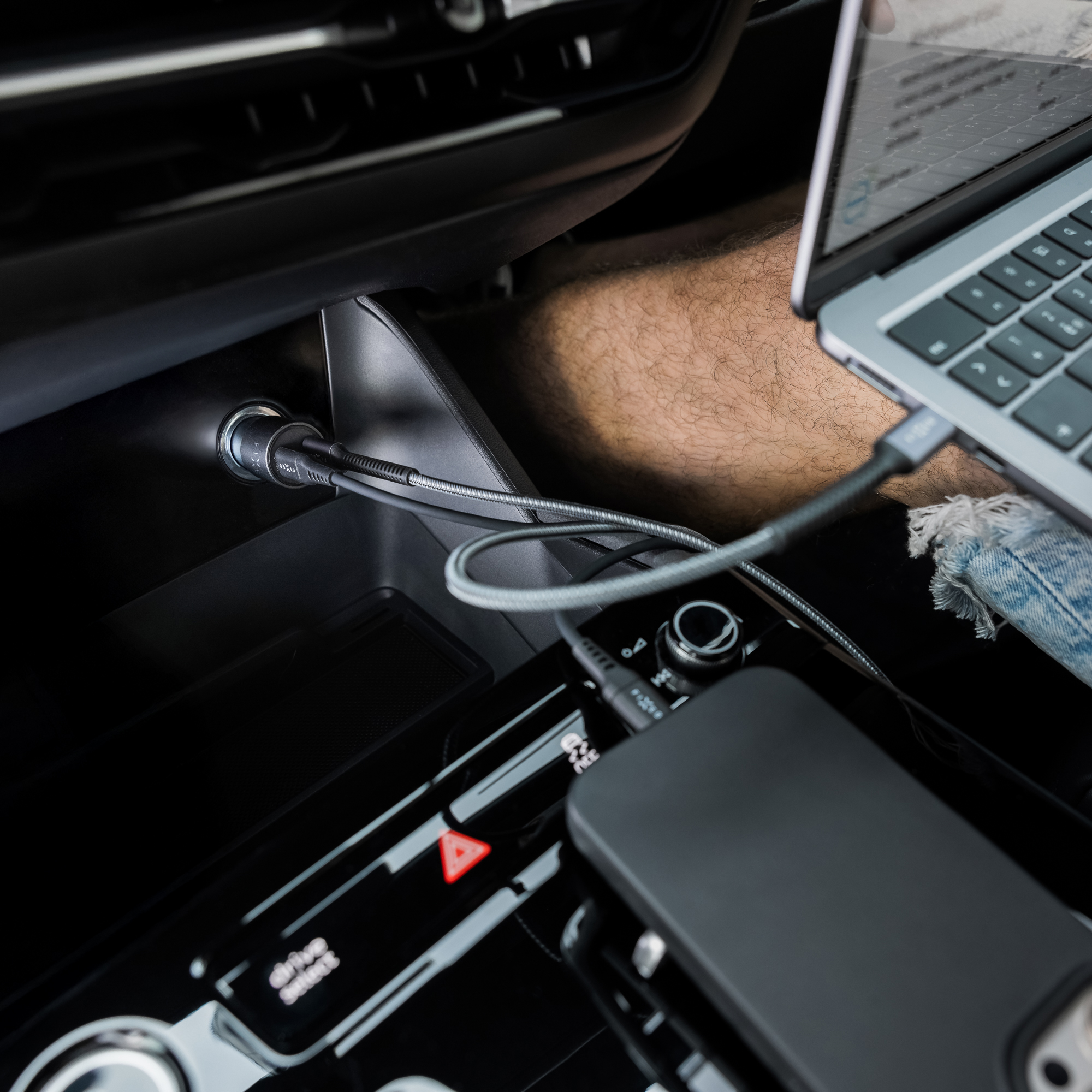 Ein Laptop, der in einem Auto aufgeladen wird. Ein Telefon und ein Autoladegerät sind ebenfalls sichtbar. Der Innenraum des Autos ist schwarz.