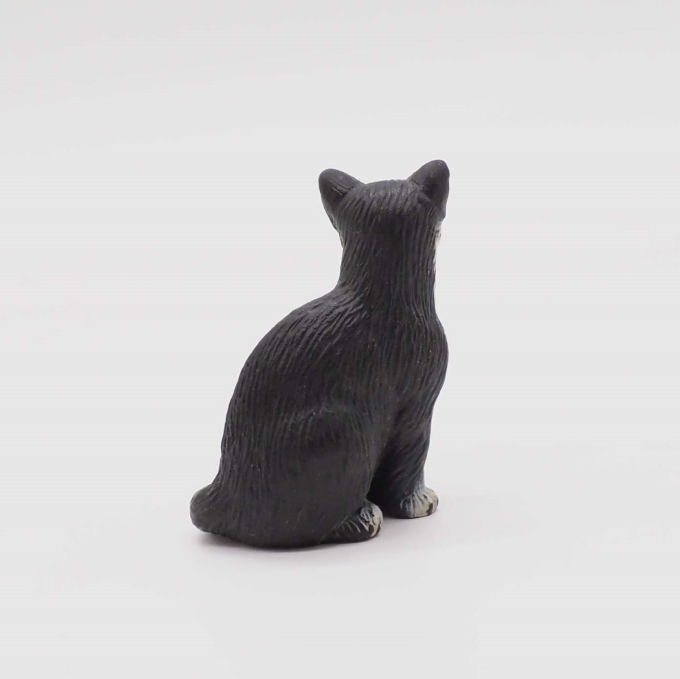 Eine schwarze Katzenfigur sitzt abgewandt auf einer weißen Oberfläche. Die Katze ist detailliert mit sichtbarer Fellstruktur und spitzen Ohren.