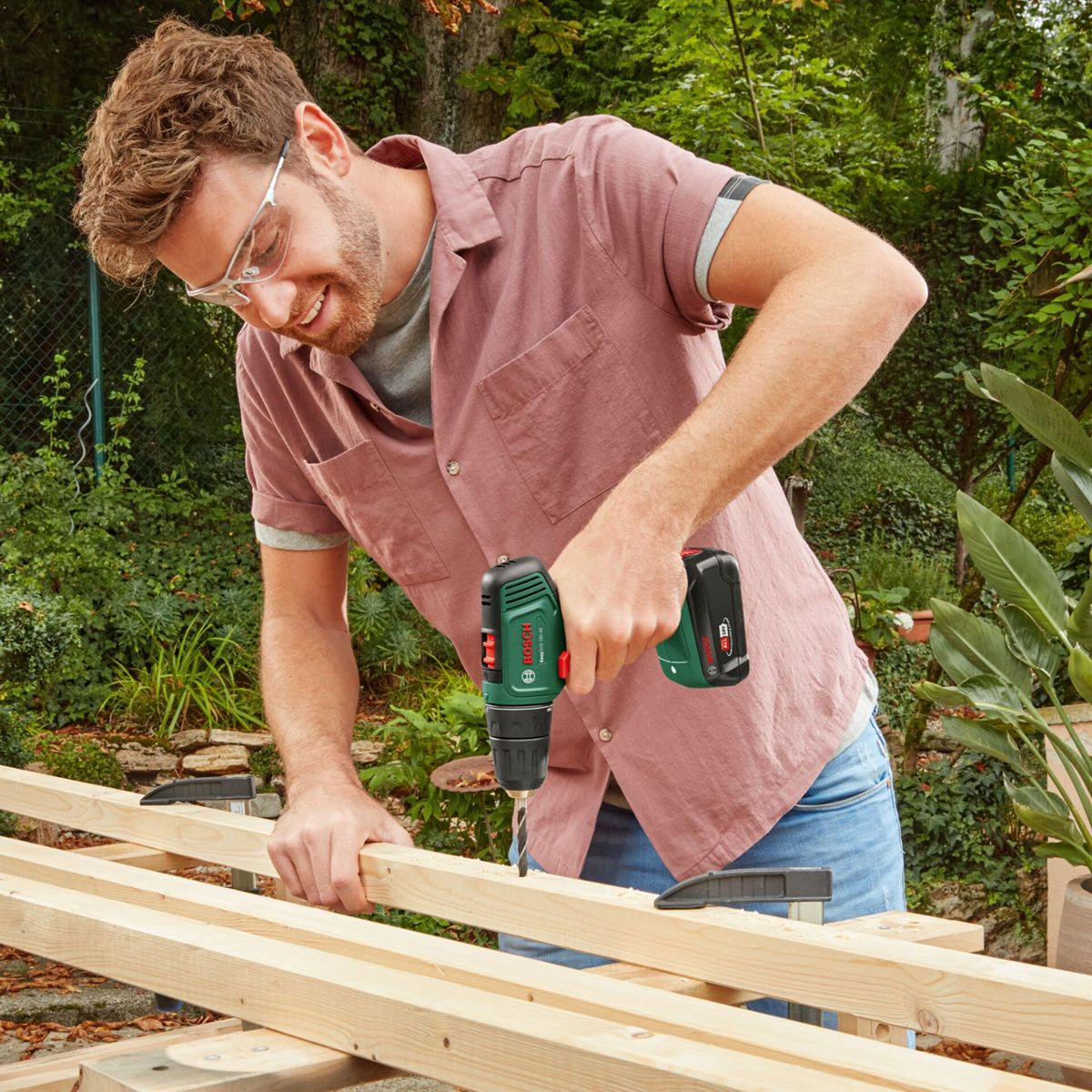 Mężczyzna wierci w drewnie na zewnątrz. Ma okulary ochronne i różową koszulę. Zielone rośliny i drewno w tle.