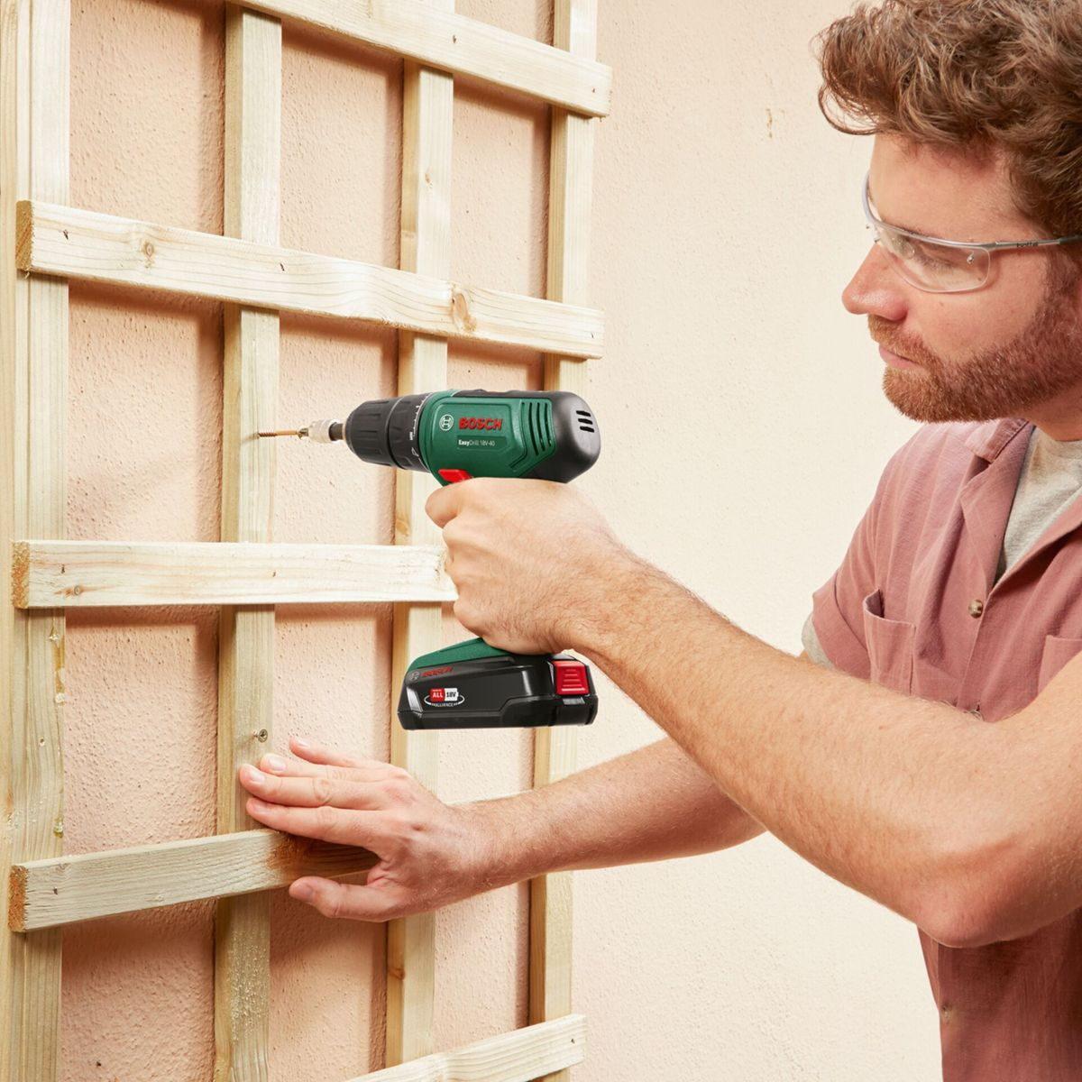 Mężczyzna wkręca śrubę w drewnianą kratę. Ma na sobie okulary ochronne i czerwoną koszulę. Zielono-czarna wiertarka. Beżowa ściana w tle.