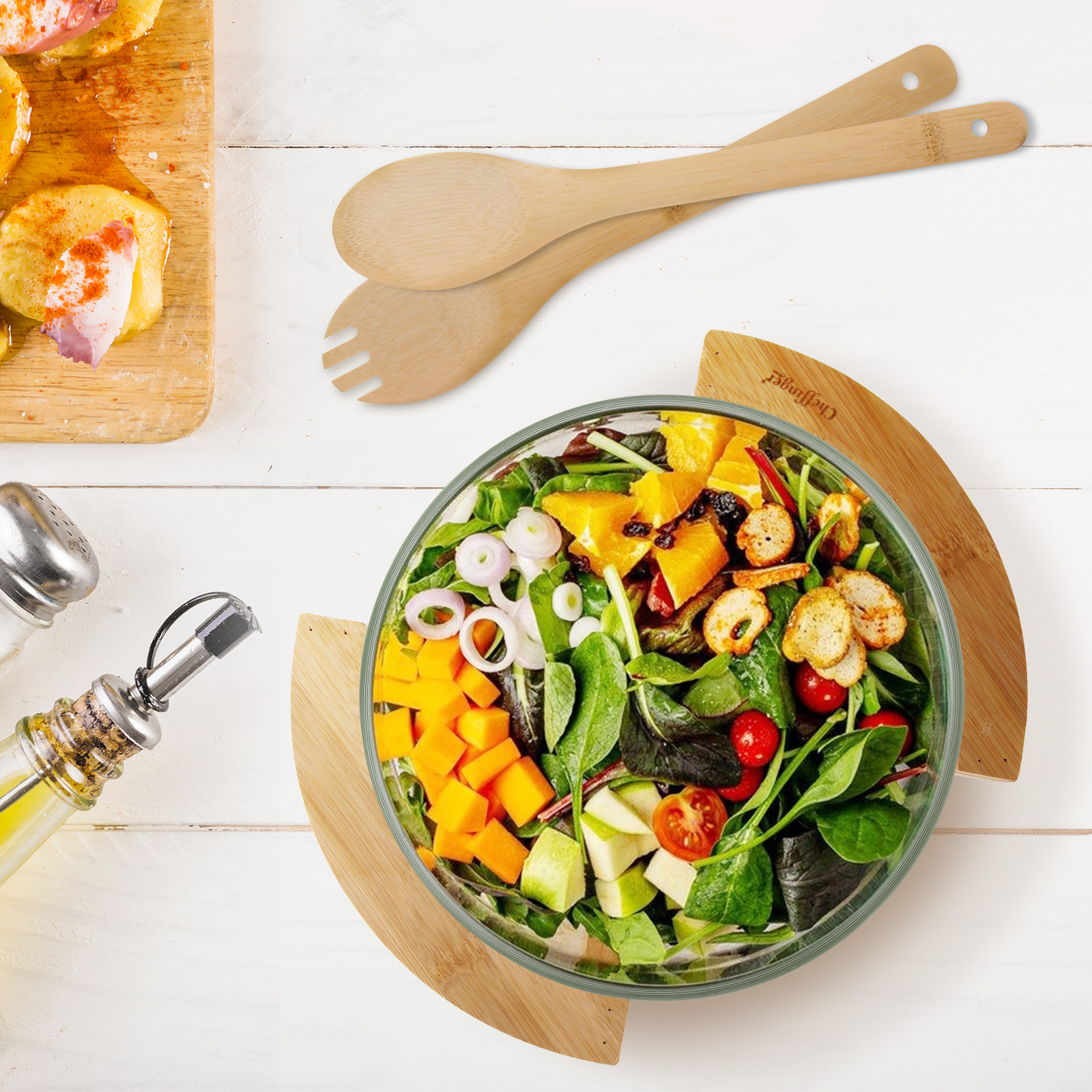 Draufsicht auf Salat in einer Glasschüssel mit Holzutensilien und anderen Lebensmitteln auf weißer Holzoberfläche.