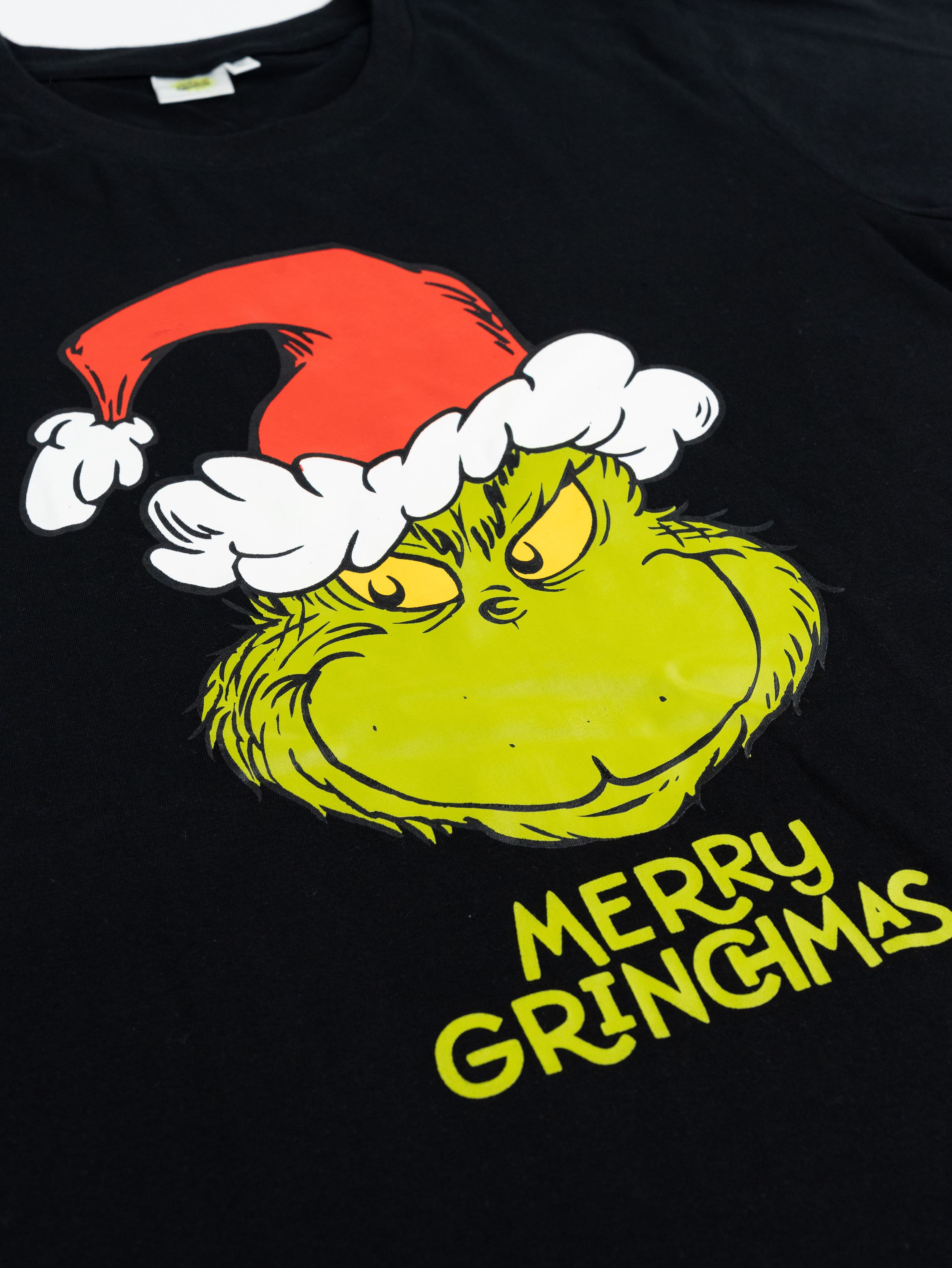Schwarzes T-Shirt mit Grinch-Gesicht und Weihnachtsmütze. Text: Merry Grinchmas. Grüner Grinch, roter Hut, weißer Besatz.