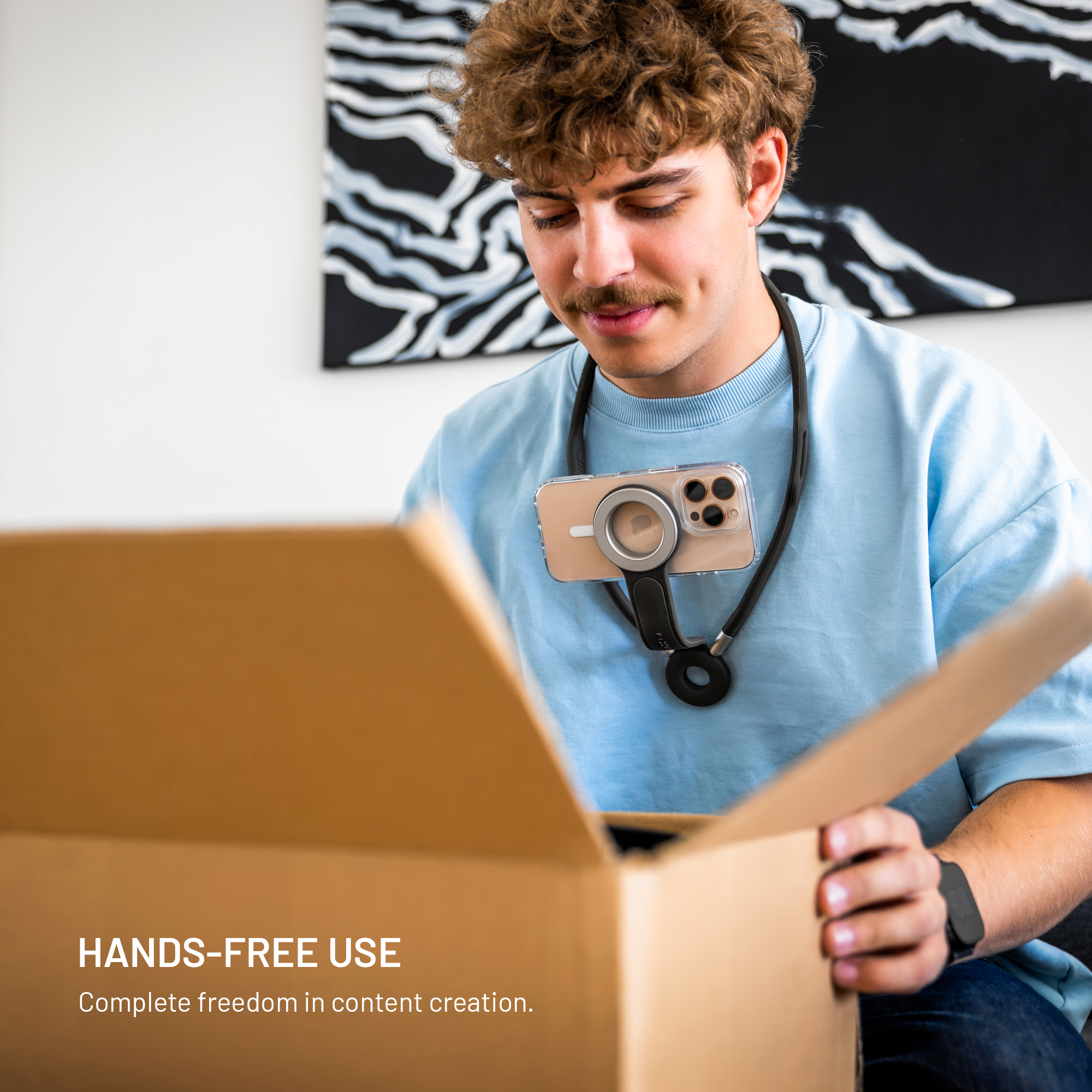 Mann mit Telefon um den Hals, öffnet eine Box. Er trägt ein blaues Hemd und hat braune Haare, Schnurrbart. Hintergrund zeigt ein Gemälde.