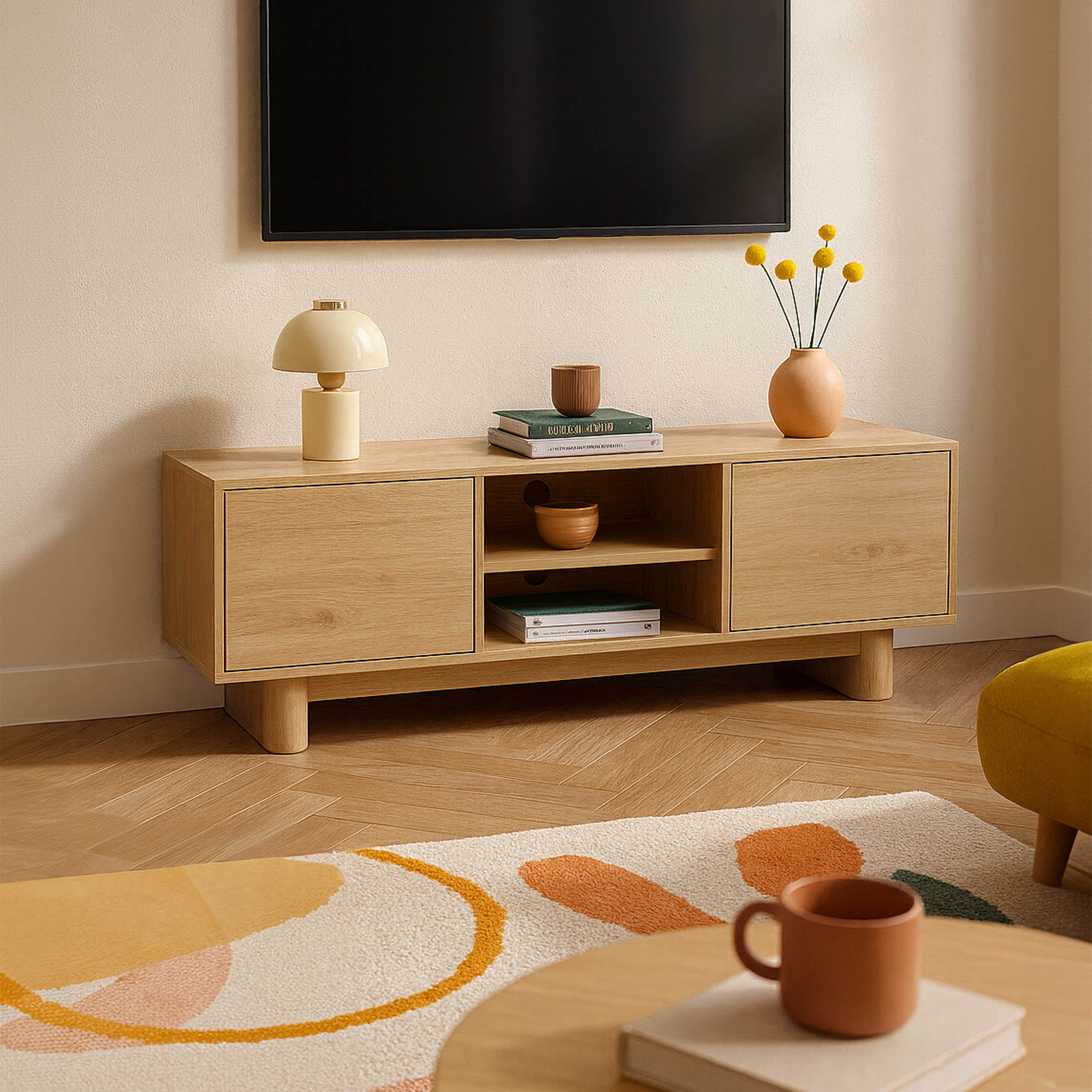 Ein Holzschrank mit einem Fernseher darüber. Auf dem Schrank befinden sich Bücher, eine Lampe und eine Vase. Ein Teppich und eine Tasse stehen davor.