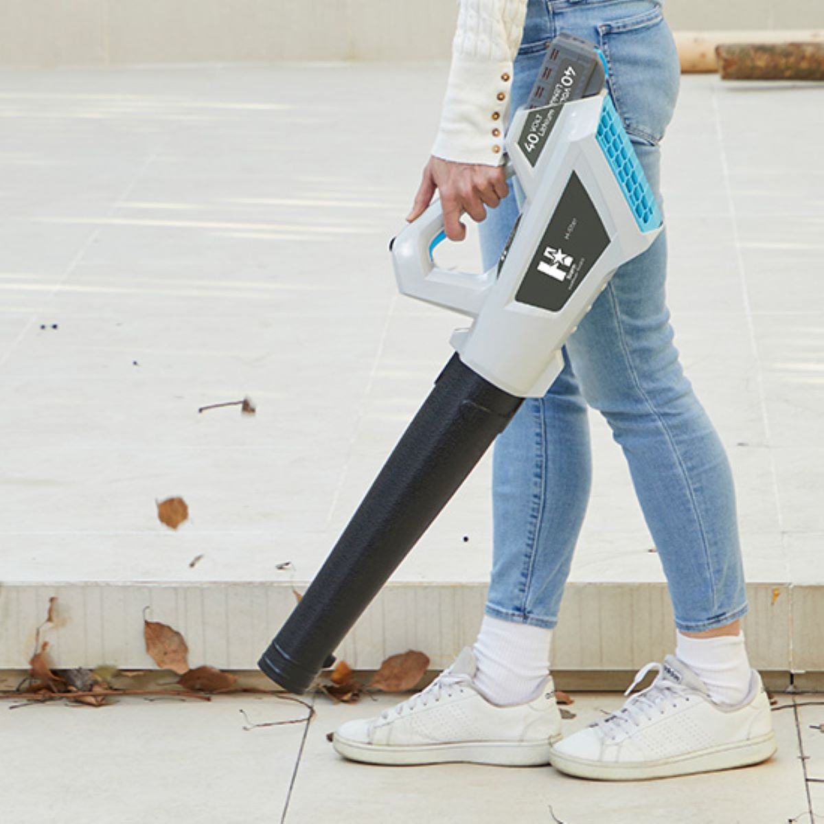 Persoon die een bladblazer vasthoudt, draagt een jeans en witte sneakers. Bruine bladeren verspreid op de grond.