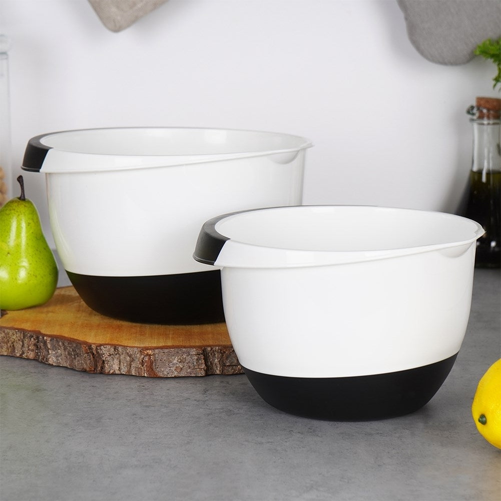 Due ciotole bianche e nere su un tagliere di legno. Una pera verde e un limone ai lati. Lo sfondo mostra oggetti da cucina.