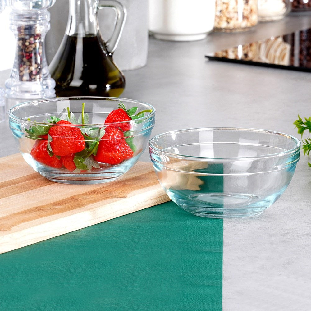 Due ciotole di vetro, una con fragole, su un tagliere di legno e una superficie verde, con oggetti da cucina sullo sfondo.