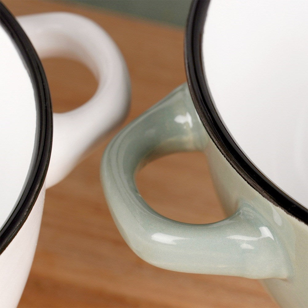 Dos tazas de esmalte blanco con bordes negros y asas verde claro. Una enfocada, la otra borrosa, sobre una superficie de madera.