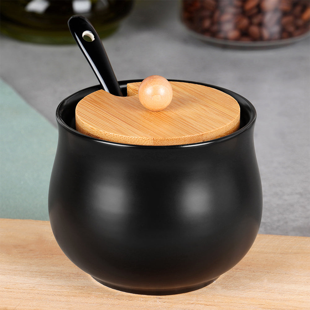 Una zuccheriera in ceramica nera con coperchio in legno e cucchiaio nero è su una superficie di legno. Lo sfondo è sfocato.