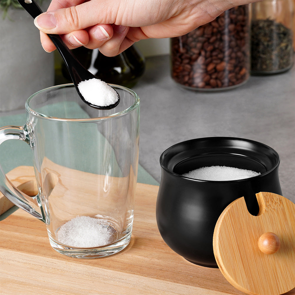 Una mano versa zucchero da un cucchiaio nero in una tazza di vetro trasparente. C'è zucchero nella tazza e un contenitore nero. Lo sfondo ha barattoli.