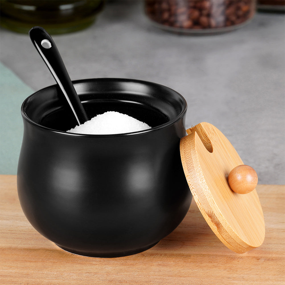 Una zuccheriera in ceramica nera con coperchio in bambù. Un cucchiaio nero è dentro la ciotola piena di zucchero bianco. Superficie di legno.
