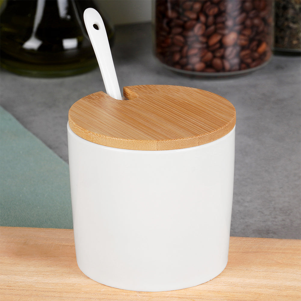 Vaso in ceramica bianca con coperchio in legno. Un cucchiaio bianco è nel vaso. Chicchi di caffè sono in un barattolo sullo sfondo.