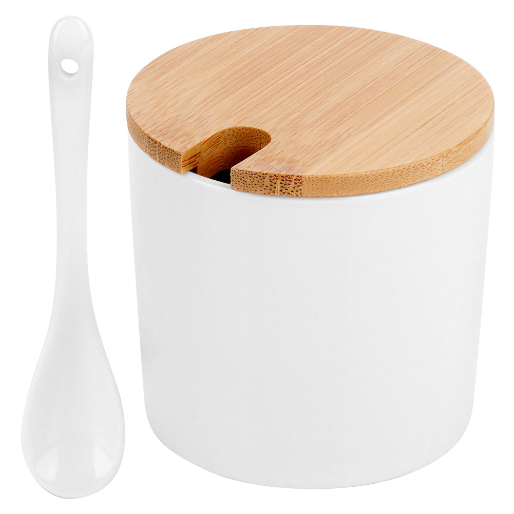 Un contenitore in ceramica bianco con coperchio in legno e cucchiaio bianco. Il contenitore è cilindrico. Il cucchiaio è accanto.