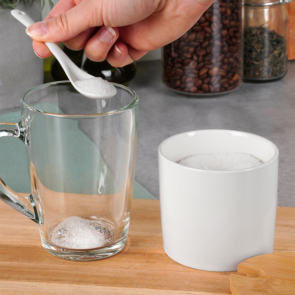 Una mano versa zucchero da un cucchiaio bianco in un bicchiere. A destra c'è una tazza bianca piena di zucchero. Vasi di chicchi di caffè sullo sfondo.