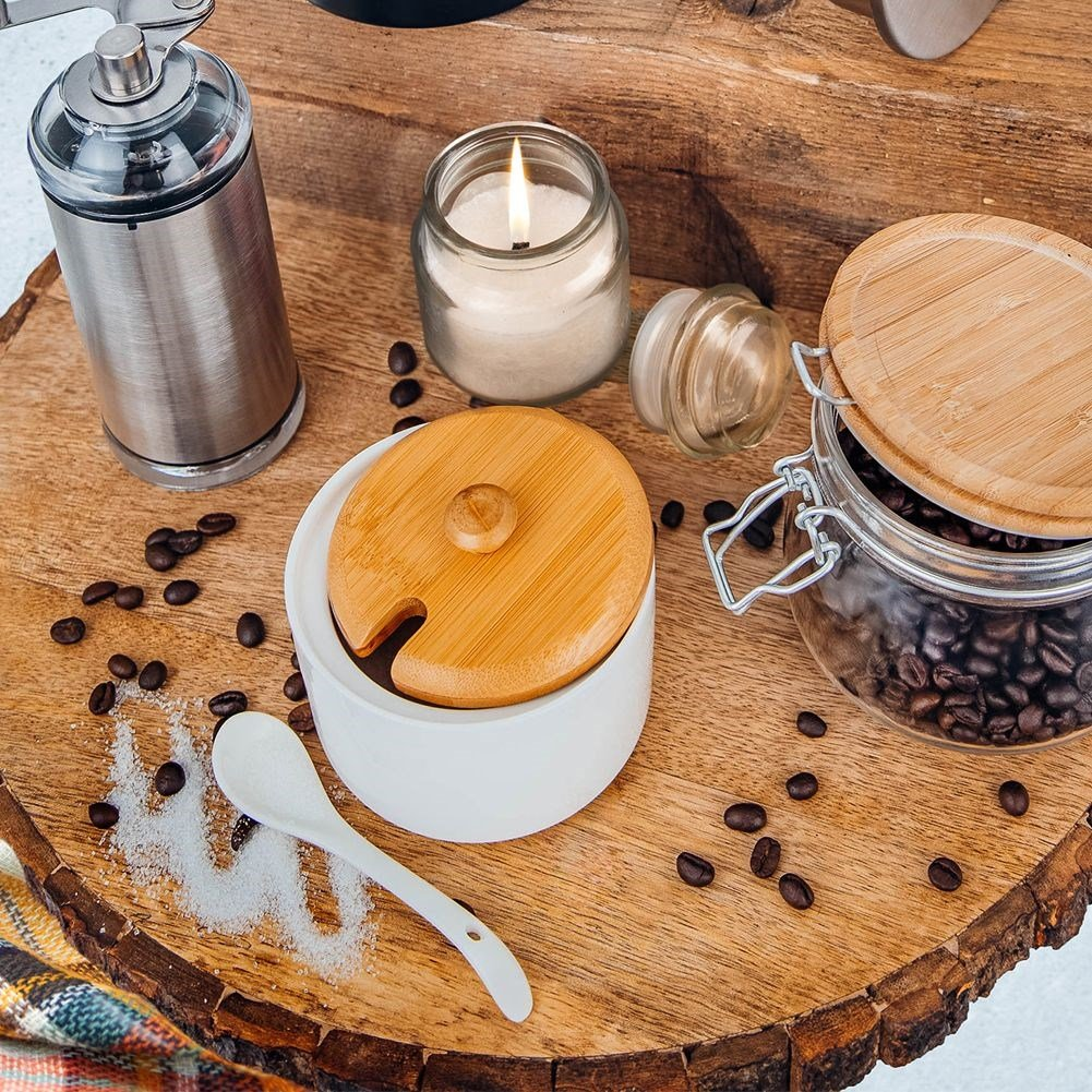 Una tavola di legno con chicchi di caffè, una candela, un barattolo con chicchi, un macinino e una zuccheriera con cucchiaio.