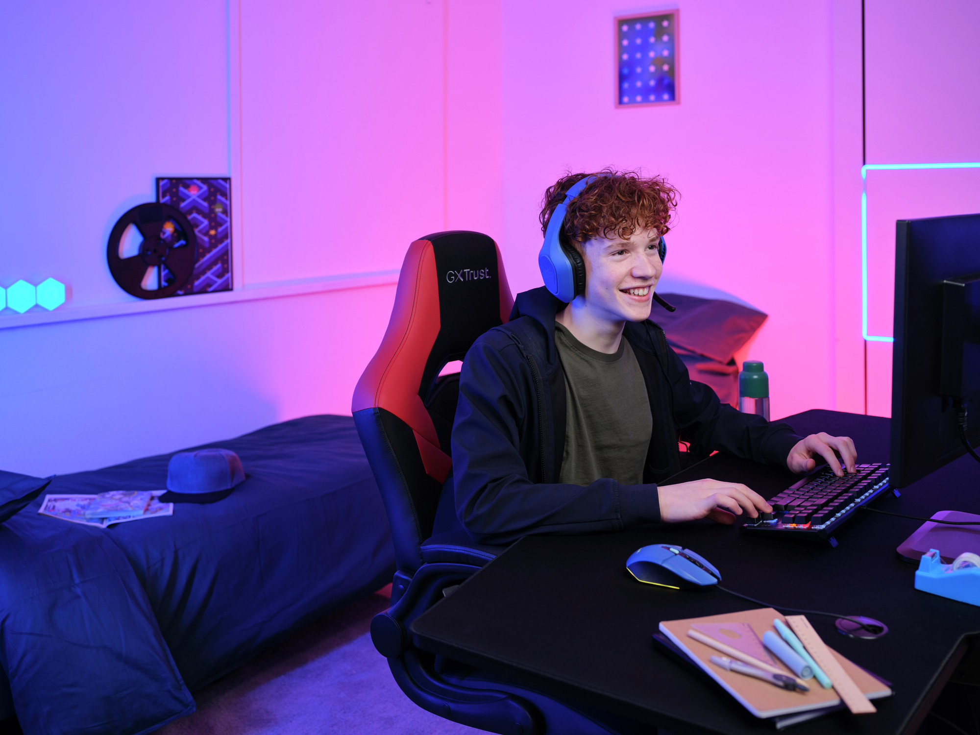 Een jonge man met een koptelefoon glimlacht terwijl hij op een computer gamet. De kamer heeft een blauw en roze kleurenschema.