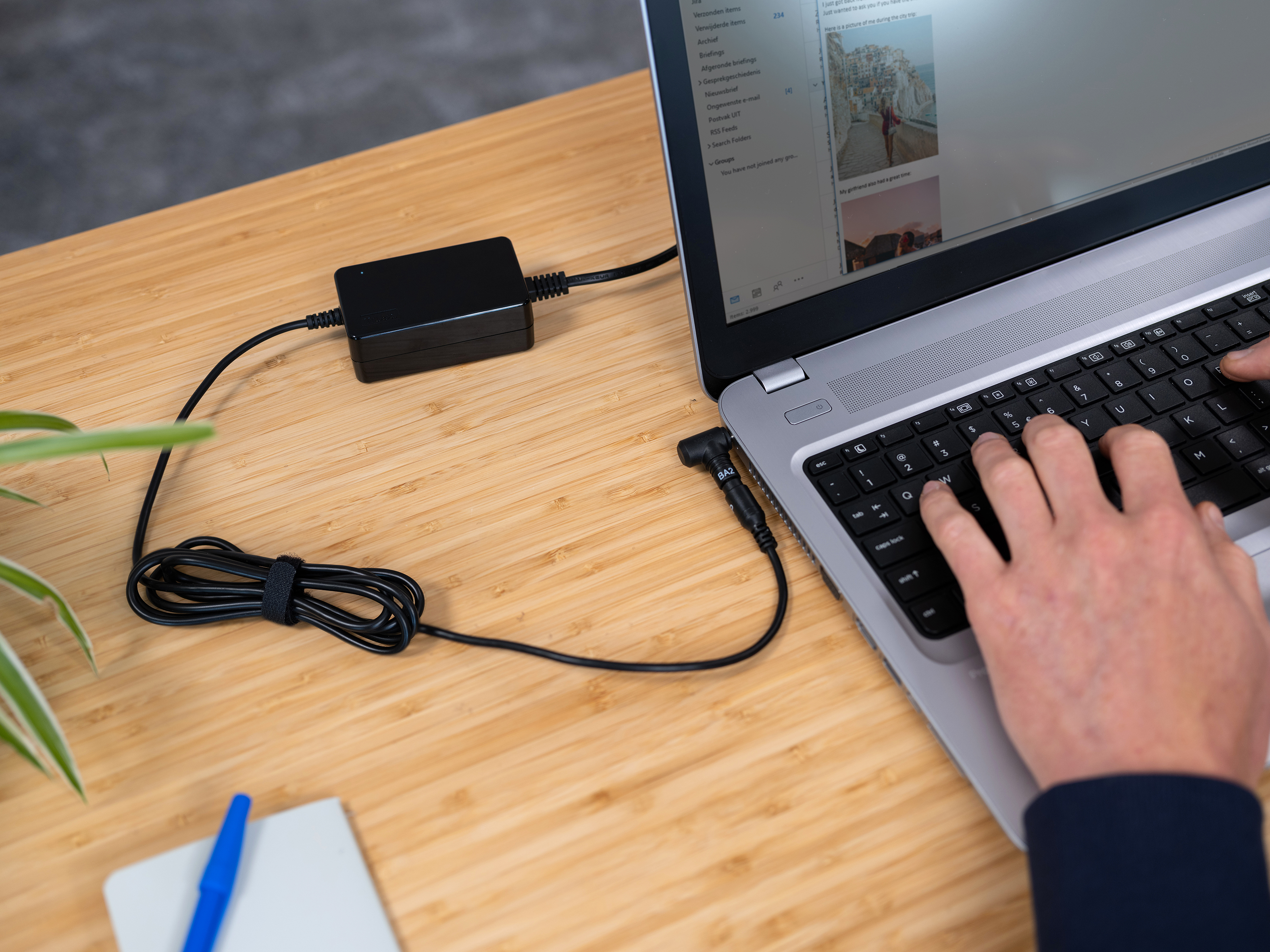 Ein Laptop auf einem Holztisch mit einem schwarzen Netzteil und Kabel. Hände, die auf der Tastatur tippen.