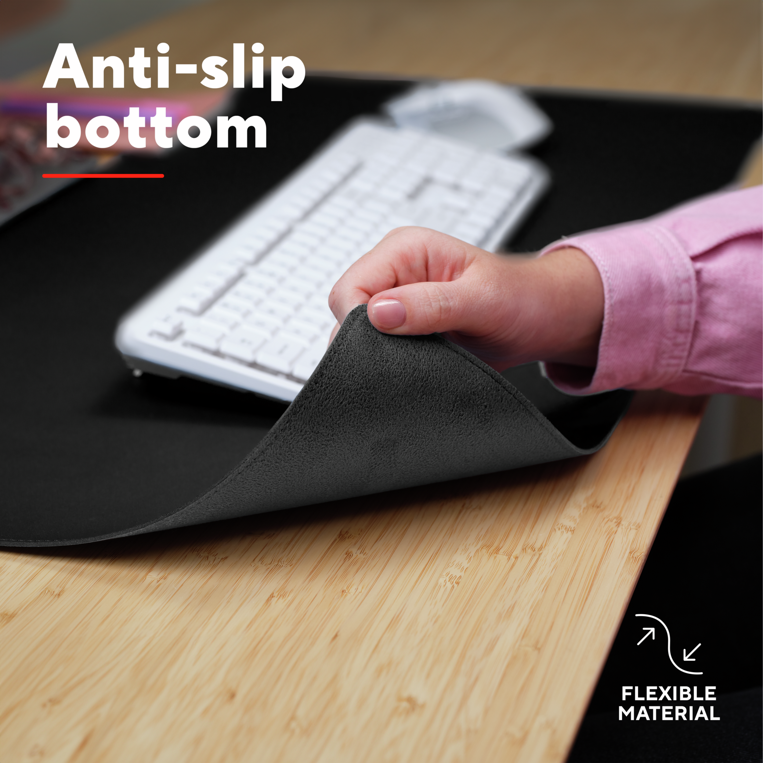 Hand holding a black anti-slip mousepad. A white keyboard is in the background, on a light wooden desk.