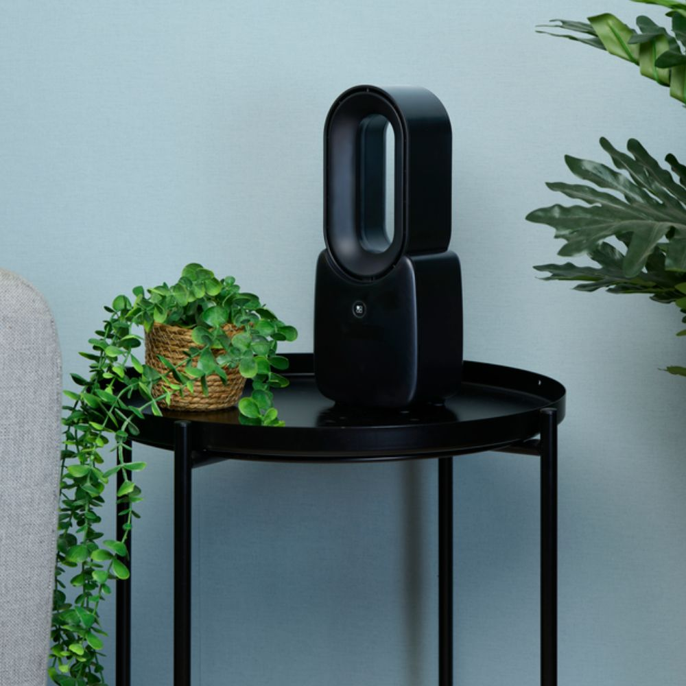 A black air purifier sits on a black table next to a plant in a woven pot. The background is a light blue wall.