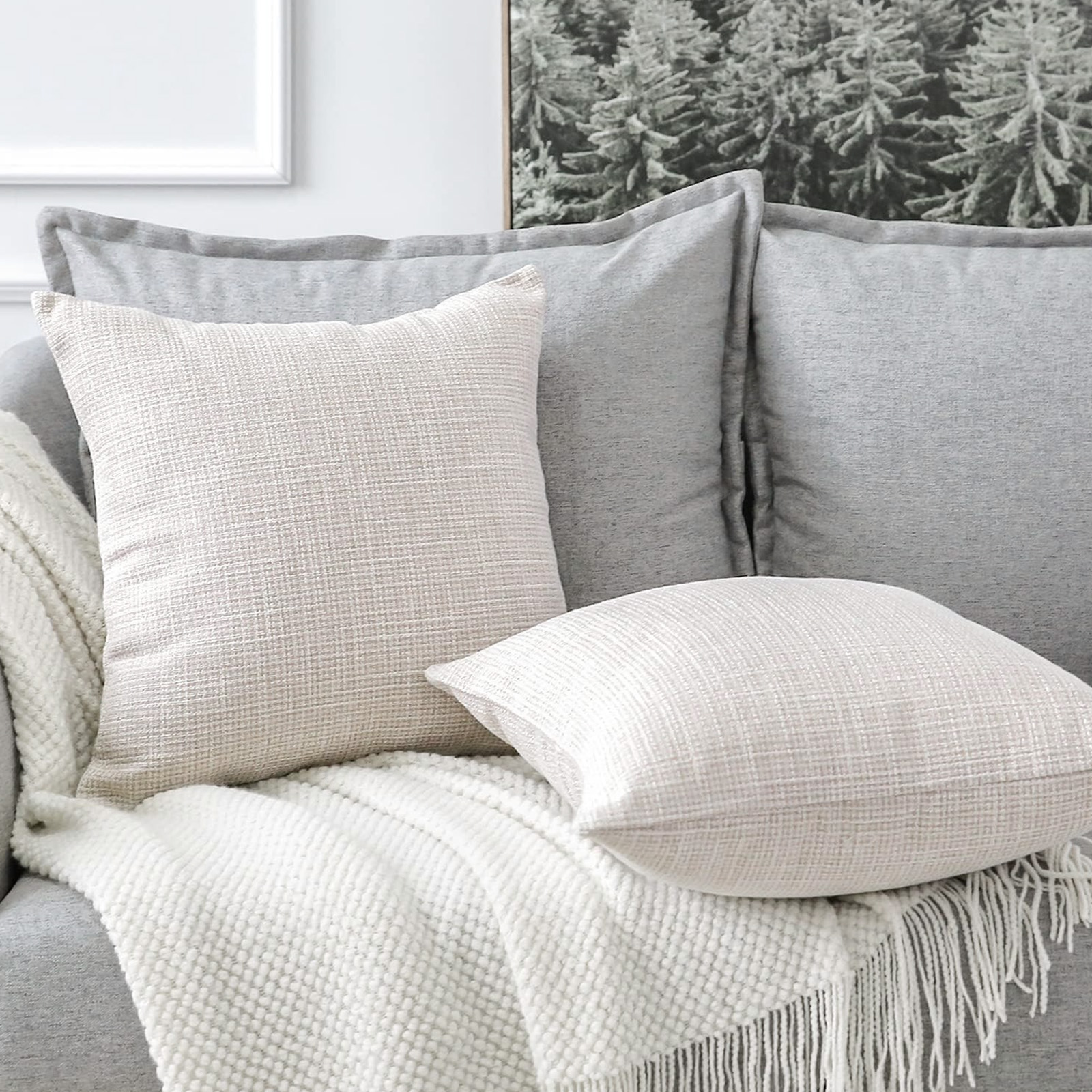 Ein Sofa mit grauen Kissen und weißen Kissen. Auf dem Sofa liegt eine weiße Decke mit Fransen. An der Wand hängt ein Waldbild.