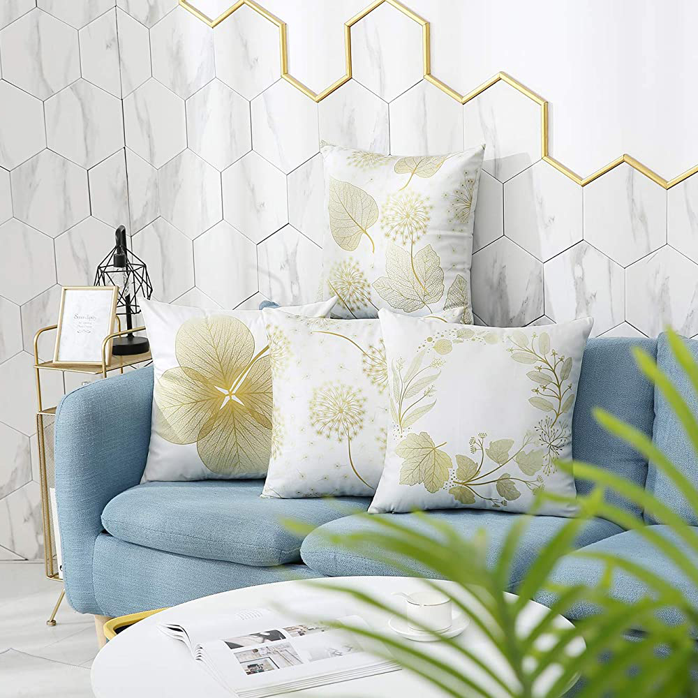 Ein blaues Sofa mit vier weißen Kissen, die mit goldenen Blättern und floralen Designs verziert sind. Ein weißer Tisch und grüne Pflanzen im Vordergrund.