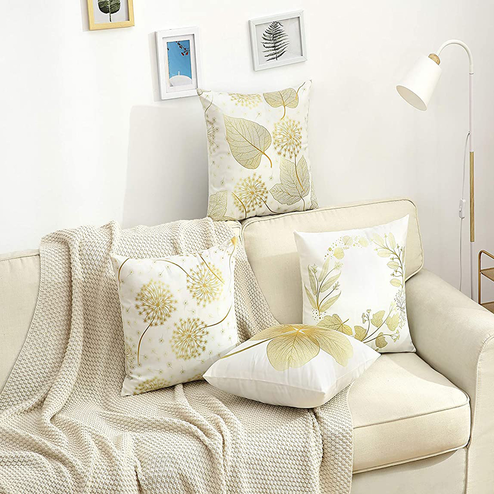 Ein beiges Sofa mit vier dekorativen Kissen mit goldenen Blättern und Löwenzahnmotiven. Eine leichte Decke und eine Lampe sind sichtbar.