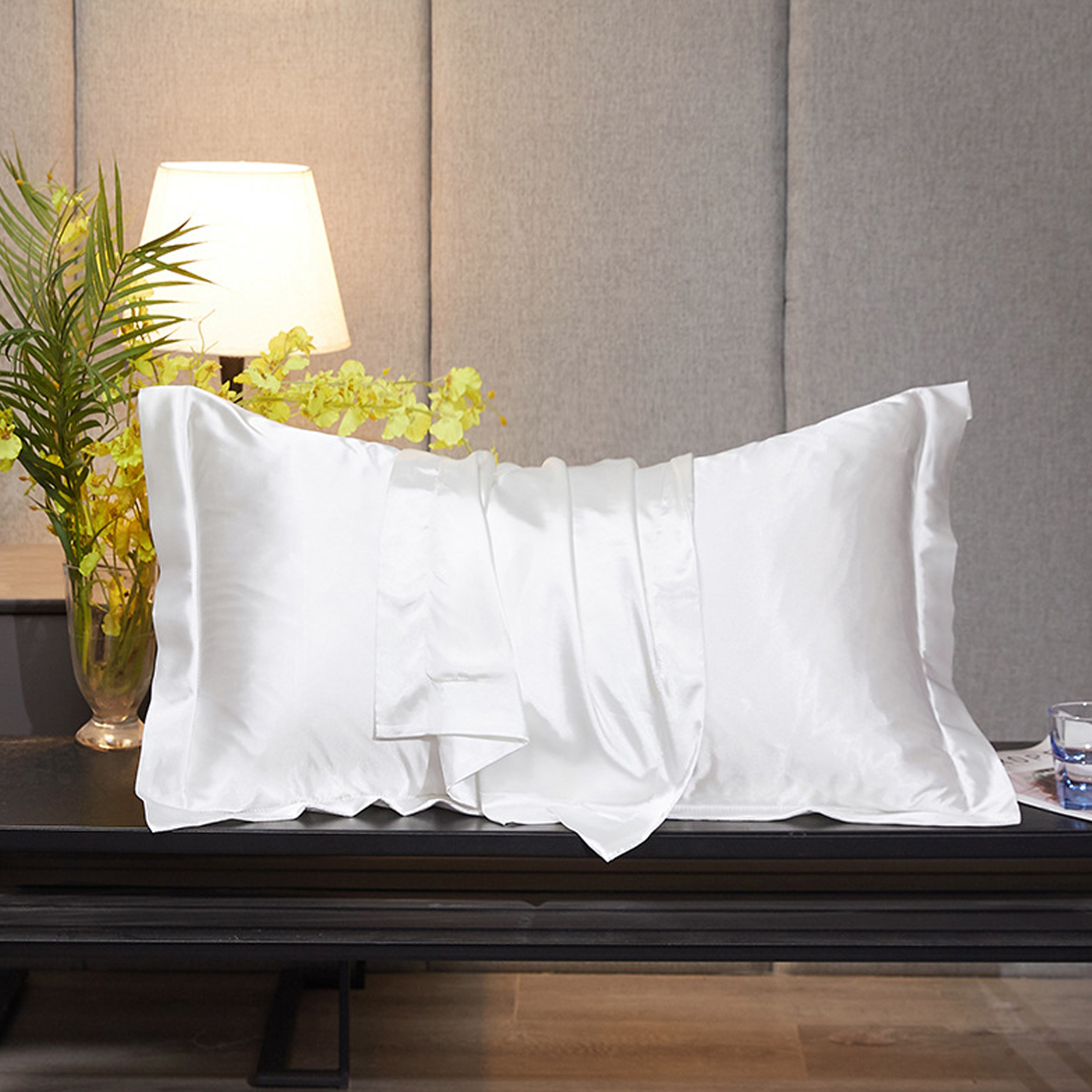 Weißes Seidenkissen auf einem schwarzen Tisch mit Lampe und Blumen. Der Hintergrund ist eine graue Wand.
