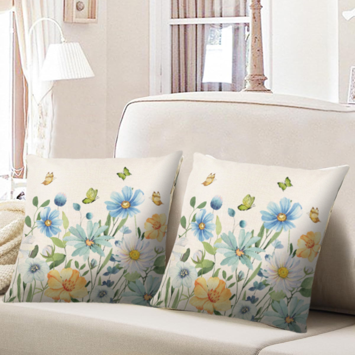 Zwei cremefarbene Kissen mit Blumendesign und Schmetterlingen liegen auf einem weißen Sofa. Im Hintergrund sind ein Fenster und eine Lampe zu sehen.