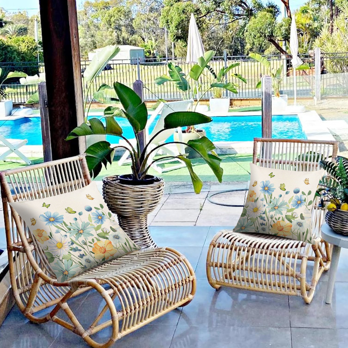 Zwei Korbstühle mit Blumenkissen stehen auf einer Terrasse mit Blick auf einen Pool. Pflanzen in Töpfen sind sichtbar.