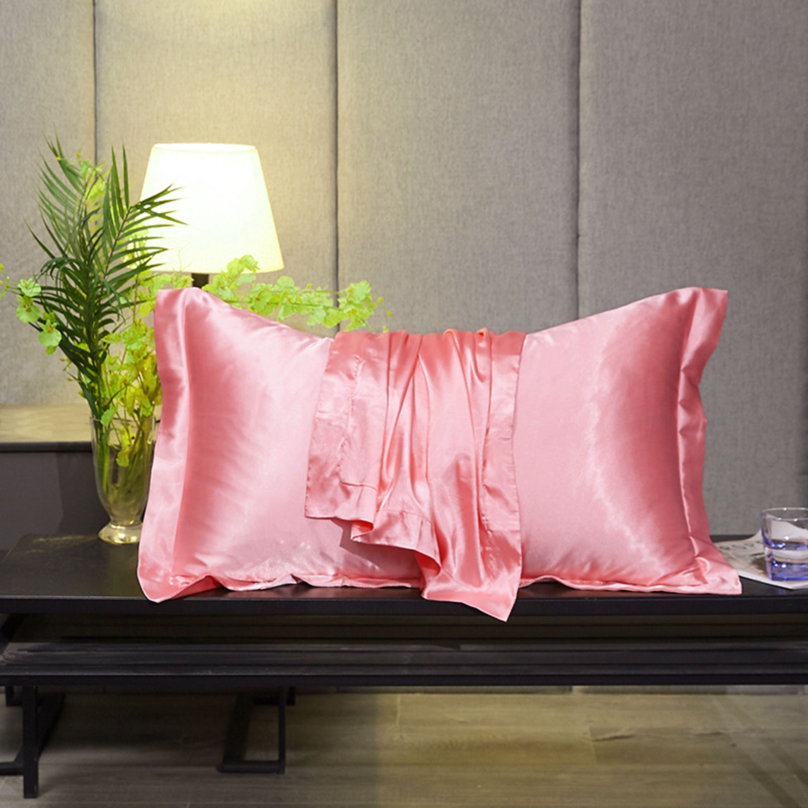 Ein rosa Seidenkissen mit drapiertem Bezug liegt auf einem schwarzen Tisch. Eine Lampe, eine Vase mit Pflanzen und ein Glas sind ebenfalls vorhanden.