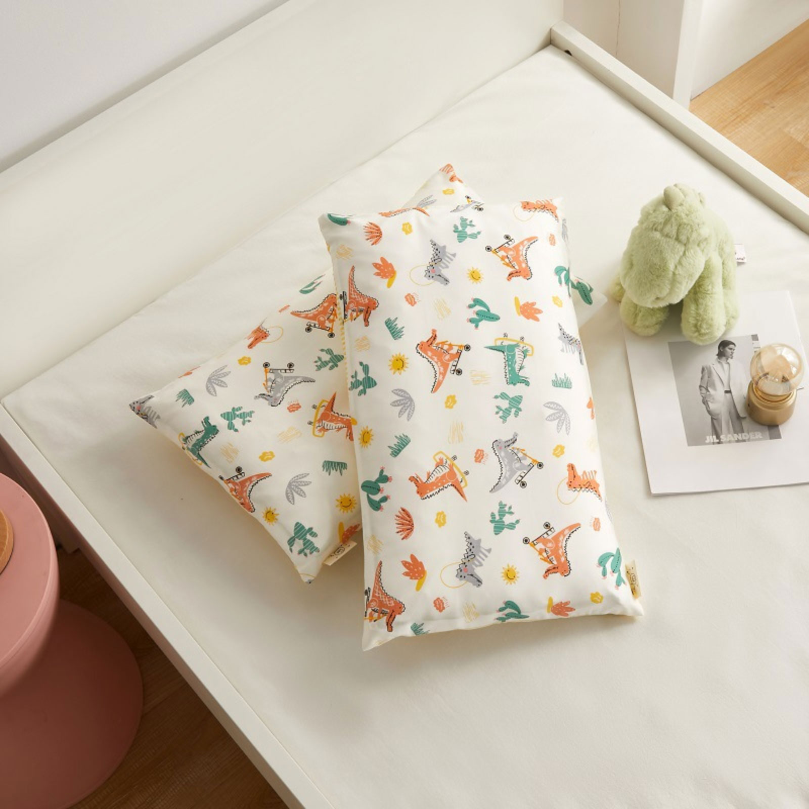 Zwei Kissen mit Dinosaurier-Muster auf einem weißen Bett. Ein grünes Stofftier und eine Zeitschrift liegen daneben.