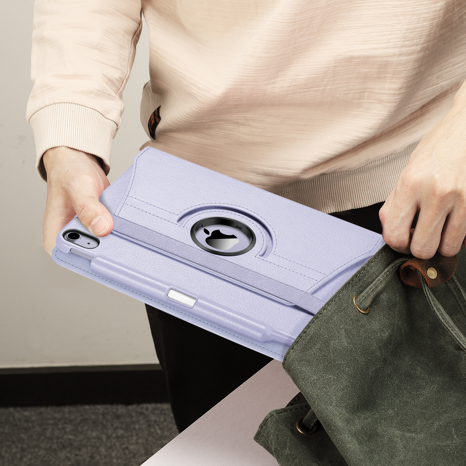 Eine Person hält eine hellviolette Tablethülle und legt sie in eine grüne Canvas-Tasche. Die Hülle hat einen Riemen und einen Stifthalter.
