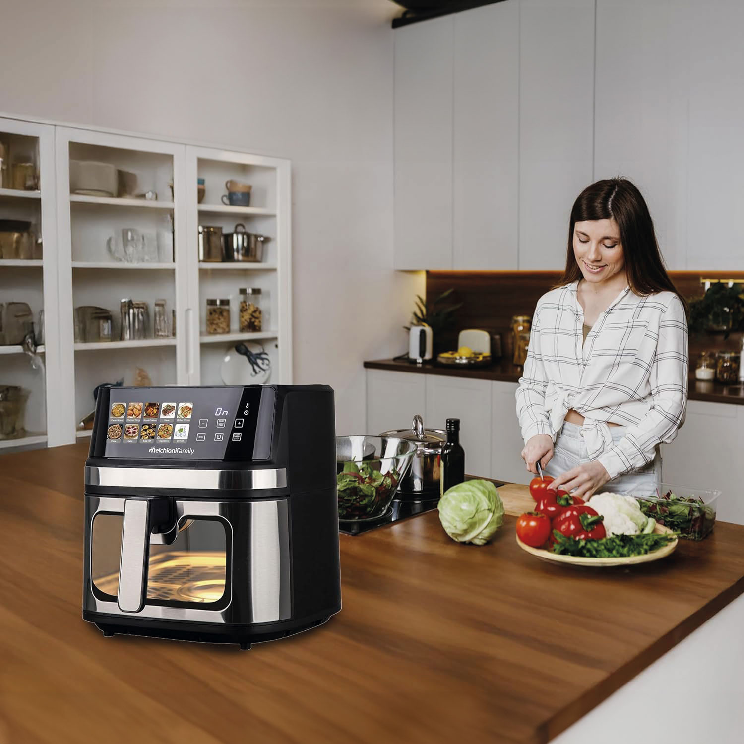 Mujer cortando verduras en una cocina. Freidora a la izquierda. Verduras en mesa de madera. Armarios de cocina atrás.
