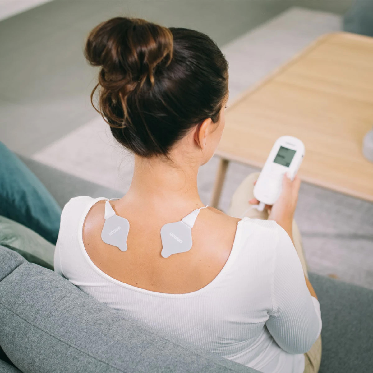 Eine Frau sitzt und benutzt ein Nackenmassagegerät. Zwei Pads sind an ihrem Rücken befestigt, und sie hält eine Fernbedienung.