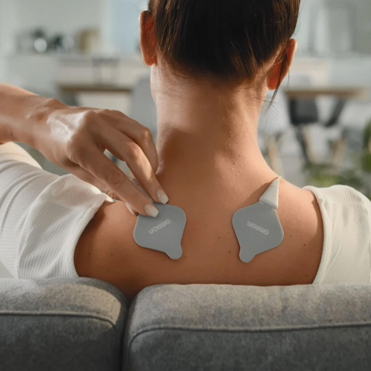 Rücken einer Frau mit Elektrotherapie-Pads. Sie sitzt und berührt ein Pad. Die Pads sind grau, mit 'Omron' beschriftet.