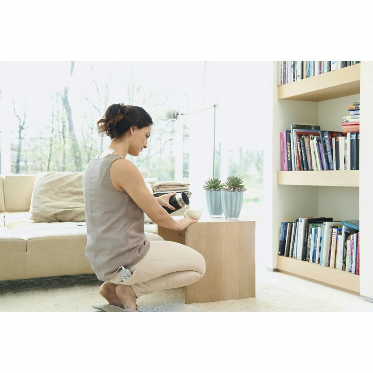 Eine Frau gießt aus einer Thermoskanne in eine Tasse. Sie steht in der Nähe eines Tisches mit Topfpflanzen, einem Bücherregal und einem Sofa.