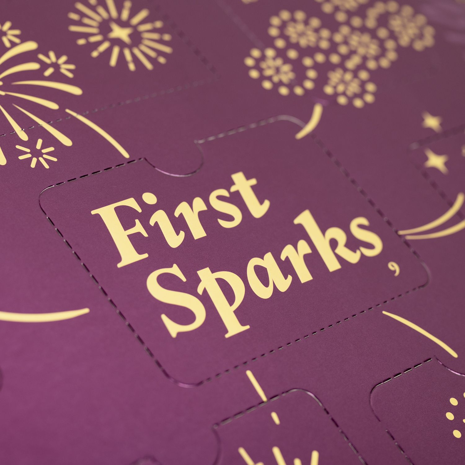 Nahaufnahme eines lila Adventskalenders. Der Text lautet 'First Sparks'. Goldene Feuerwerks- und Sternmuster sind vorhanden.