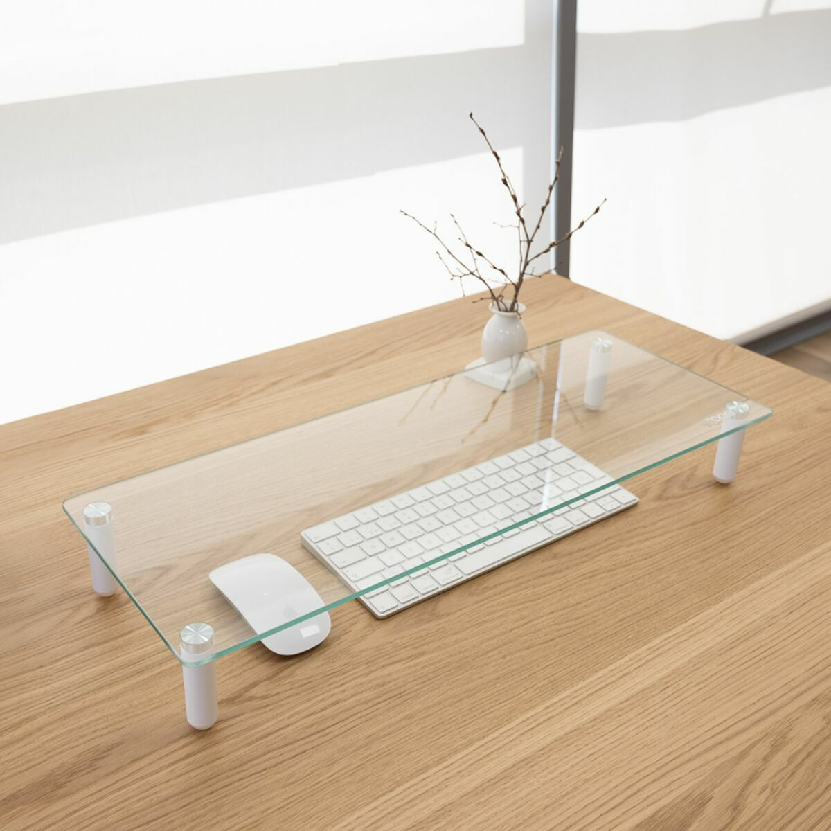 Ein Monitorständer aus Glas auf einem Holzschreibtisch hält eine Tastatur, eine Maus und eine Vase mit Zweigen. Im Hintergrund ist ein Fenster.