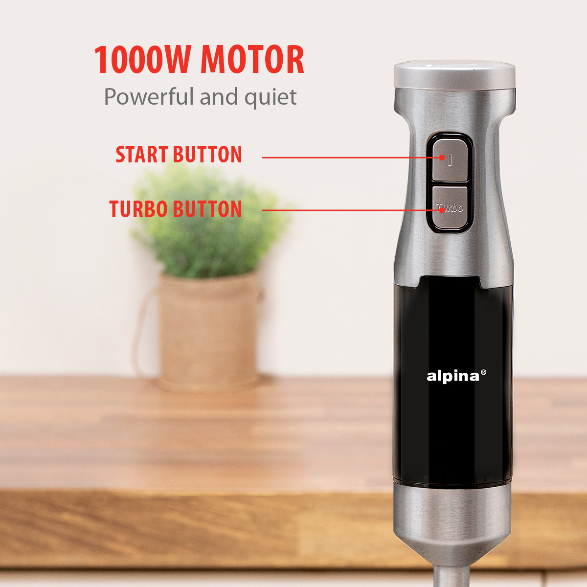 Een zilver-zwarte staafmixer met start- en turboknop staat op een houten oppervlak. Een plant in een pot staat op de achtergrond.