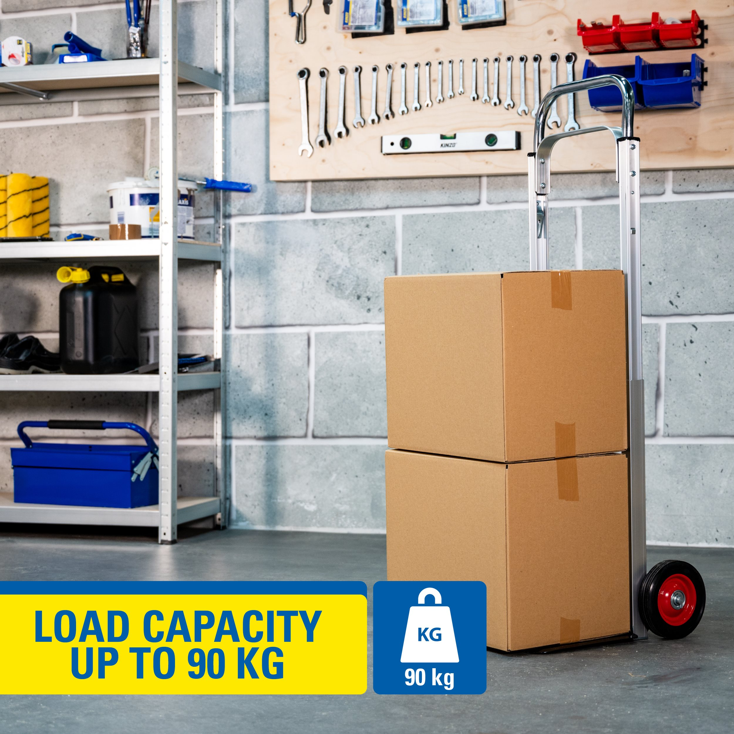 A hand truck carrying two cardboard boxes. The image shows tools and shelves in the background. Load capacity up to 90 kg.