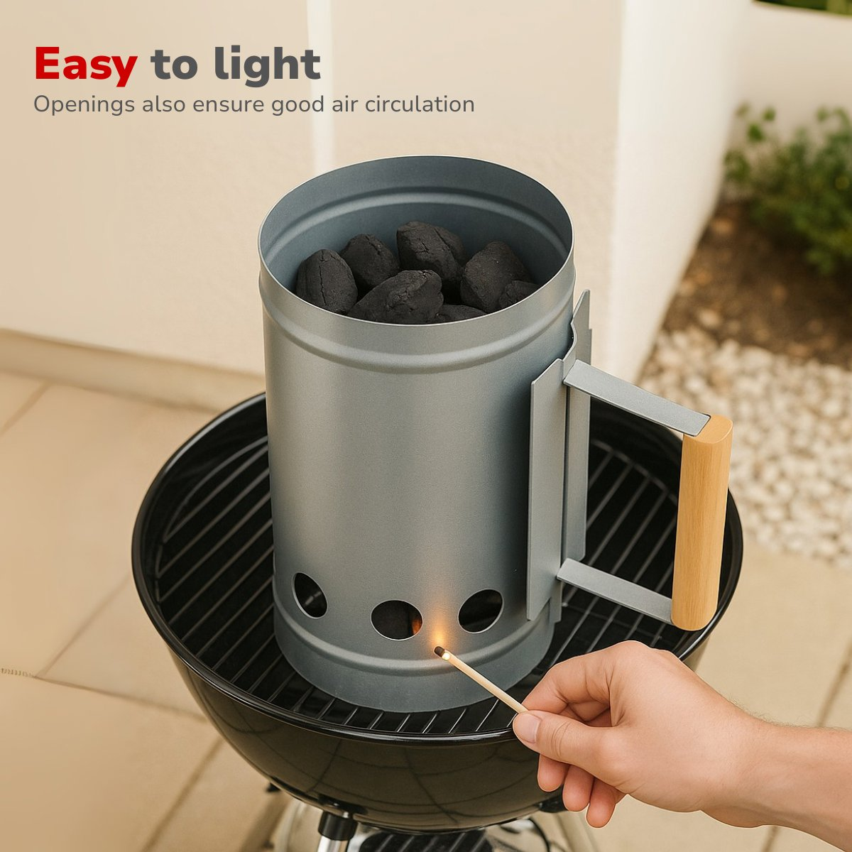 A charcoal chimney starter with lit match igniting charcoal briquettes inside, placed on a black grill. The handle is wooden.