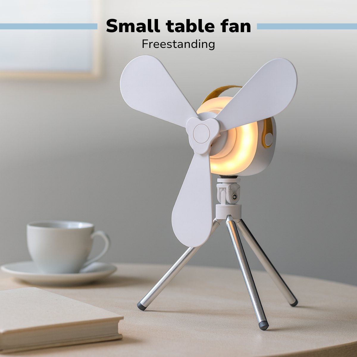 Un petit ventilateur de table blanc avec trois pales, une lumière jaune et un trépied argenté, sur une table en bois. Une tasse et un livre sont visibles.