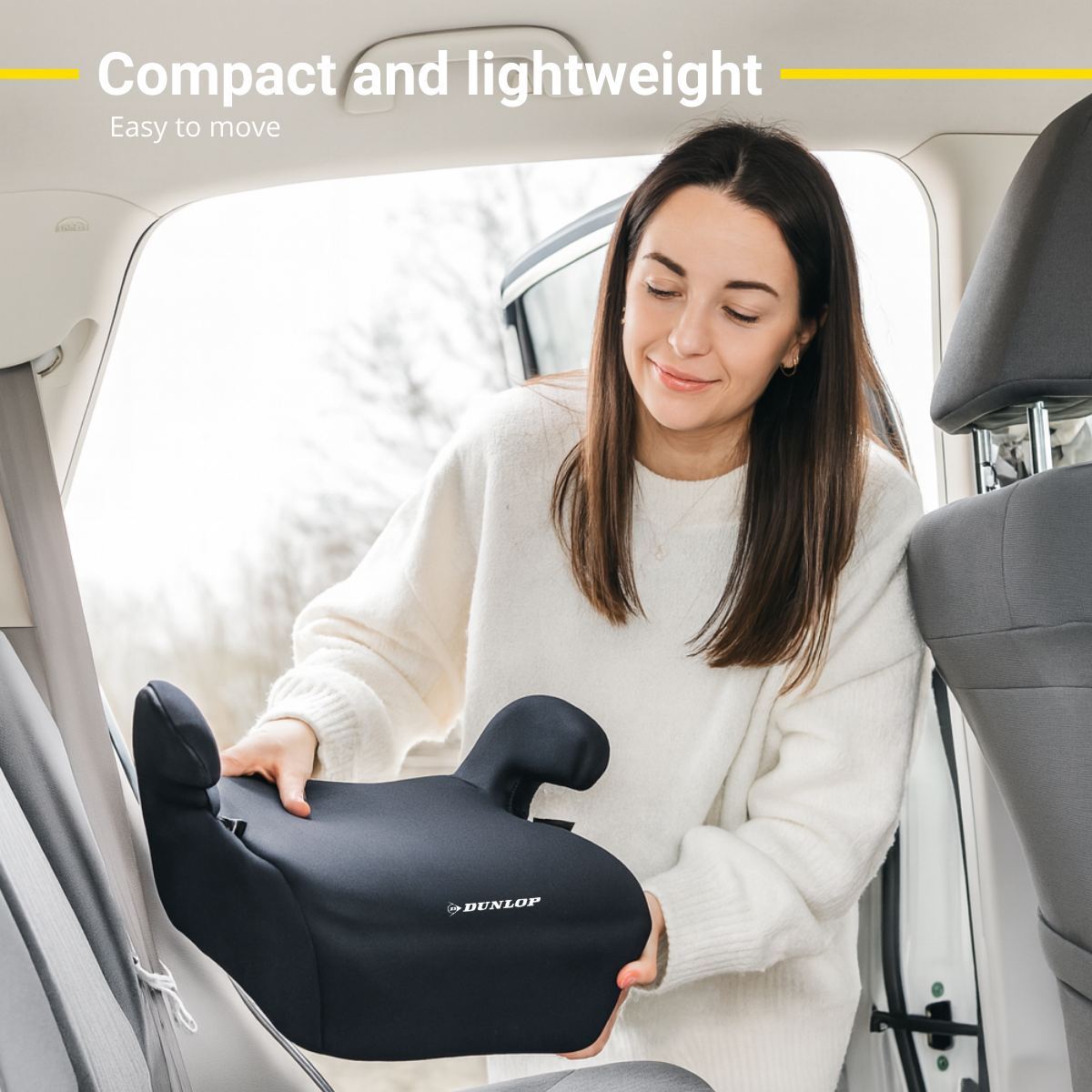 Een vrouw in een auto houdt een zwart kinderzitje vast. Het zitje heeft het Dunlop logo. Ze glimlacht. Het interieur van de auto is grijs en wit.